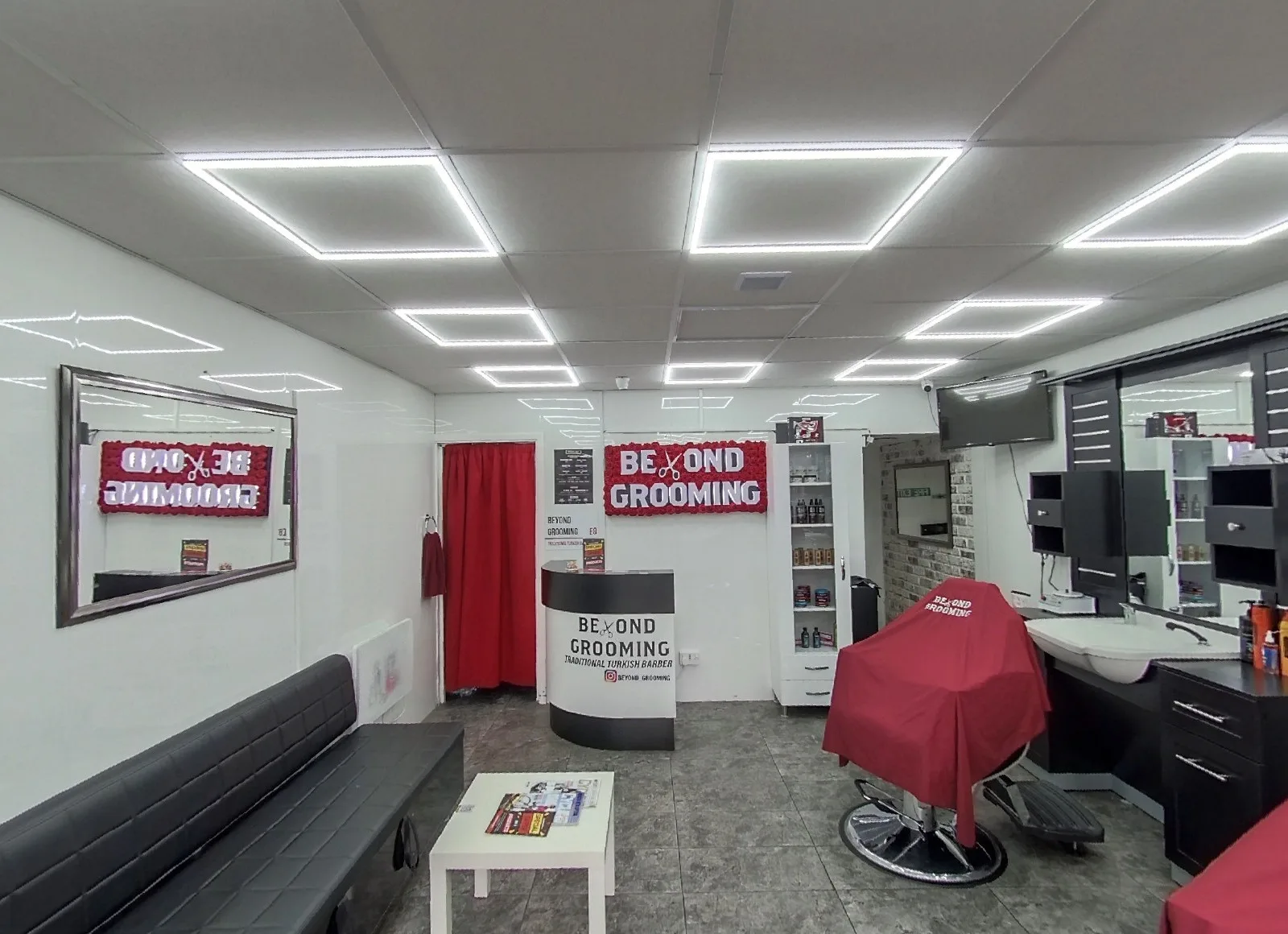 Inside Beyond Grooming: a red-caped barber chair, the red floral Beyond Grooming wall sign, reception counter, waiting bench and LED-panel ceiling