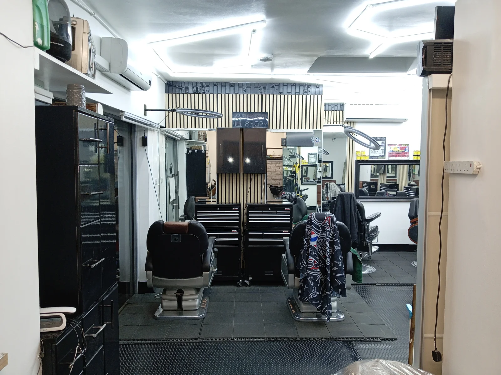 Inside Clipmason: black tool-chest barber stations, timber slat wall, ring lights, tiled floor and a patterned cape draped over an empty chair