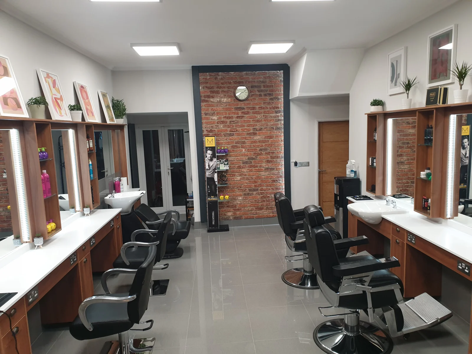 Inside Coppertones: walnut-framed mirrors with LED strips, white counters and wash basins, black salon and barber chairs, and a brick feature wall with a clock
