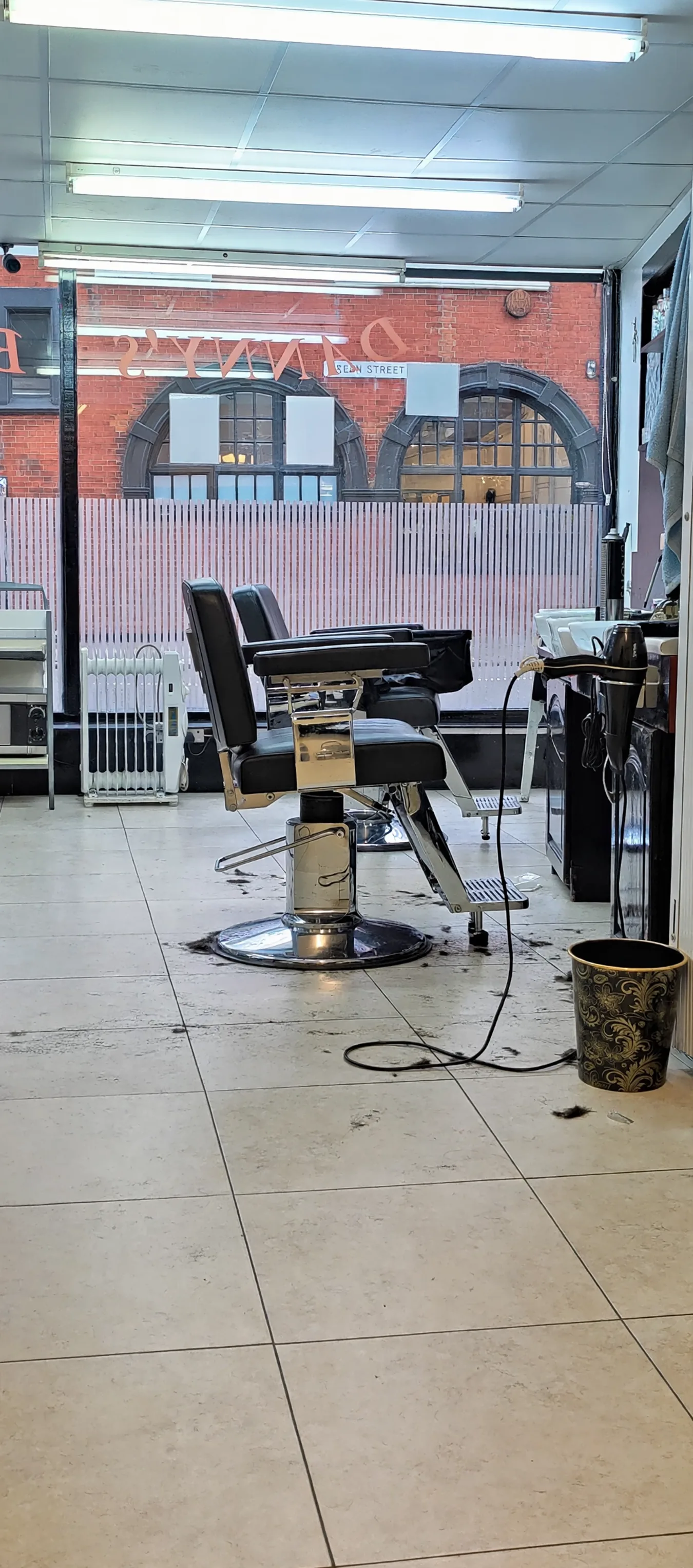 Inside Danny's Barber: empty chrome barber chairs on a cream tiled floor, with The Boleyn pub's arched windows visible through the glass front