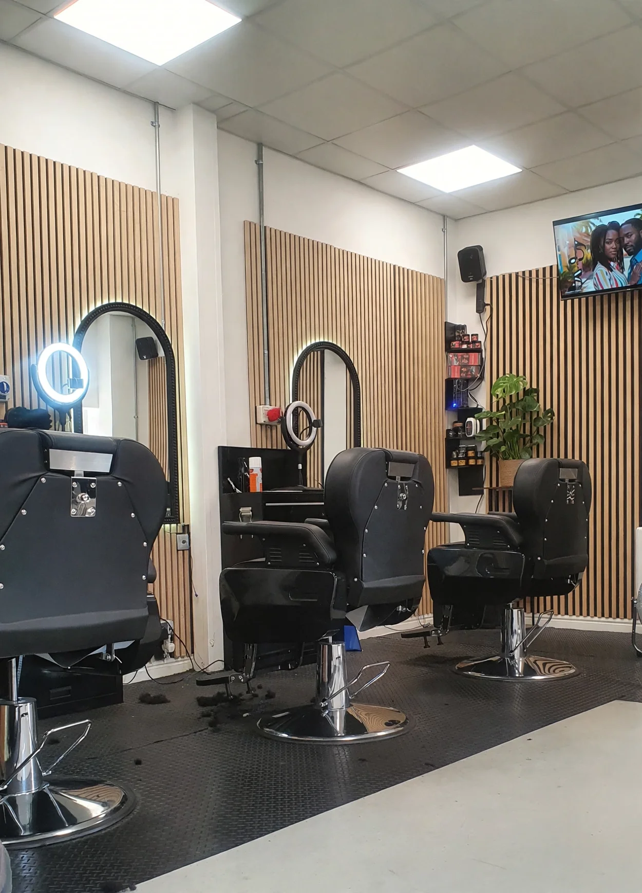 Inside Mullings Cutting Edge: light wood slat walls, arched mirrors with ring lights, three black barber chairs and a monstera plant