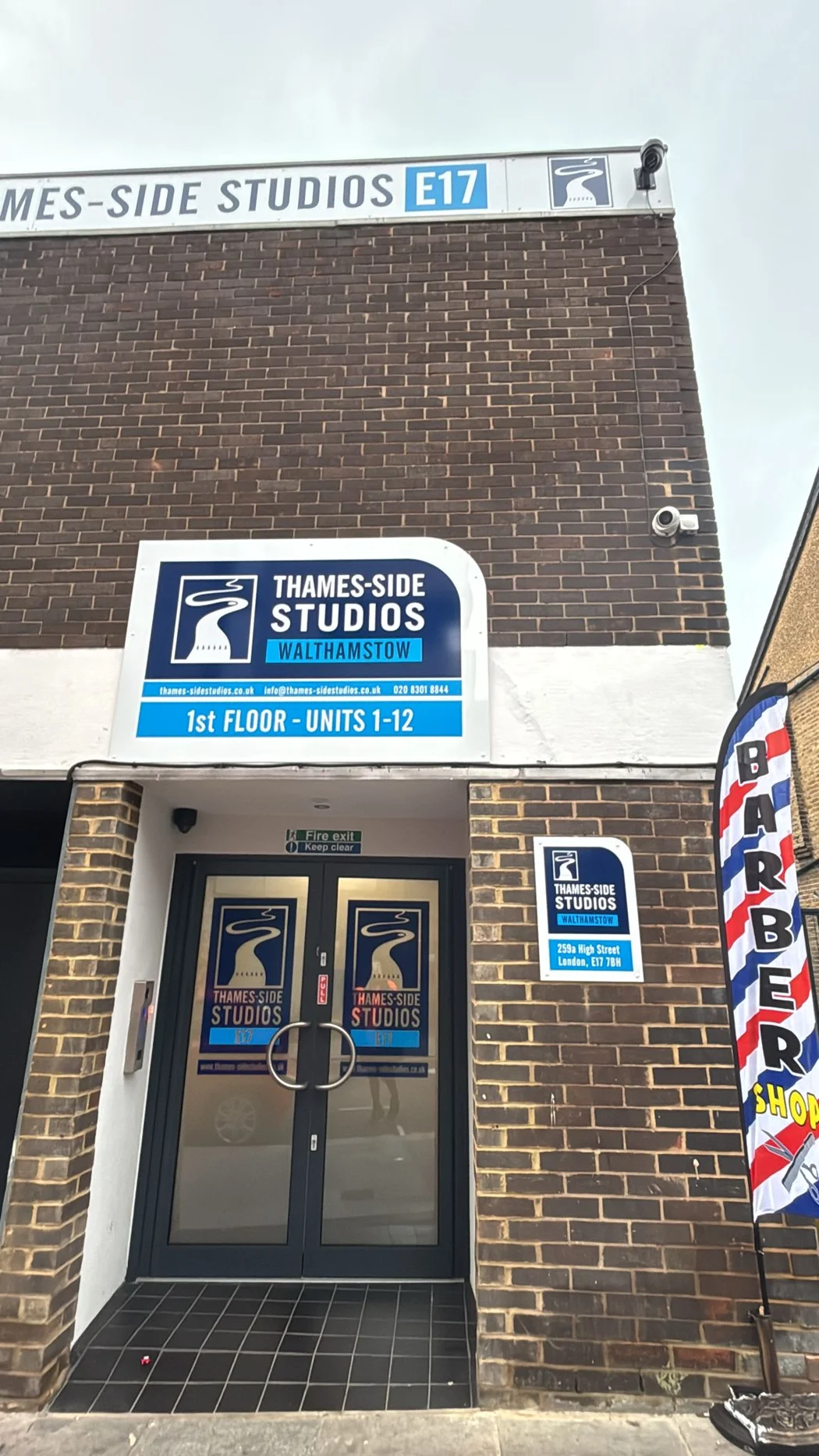 The Thames-Side Studios entrance on Walthamstow High Street: brick facade, studio signage, the 259a High Street plaque and Mullings' BARBER SHOP feather flag by the door