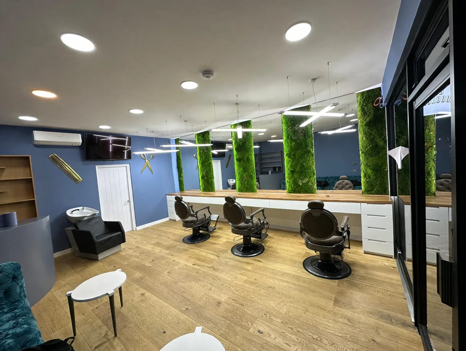 Inside Premium Barber: blue walls, moss-covered columns, backlit mirrors, X-shaped lights and three empty barber chairs
