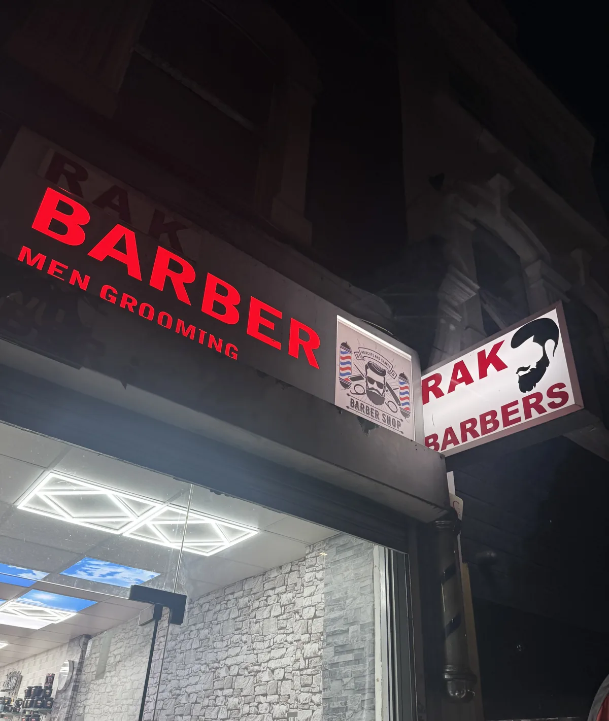 RAK Barbers at night: red neon RAK Barber Men Grooming fascia and the projecting RAK Barbers sign with bearded-man logo