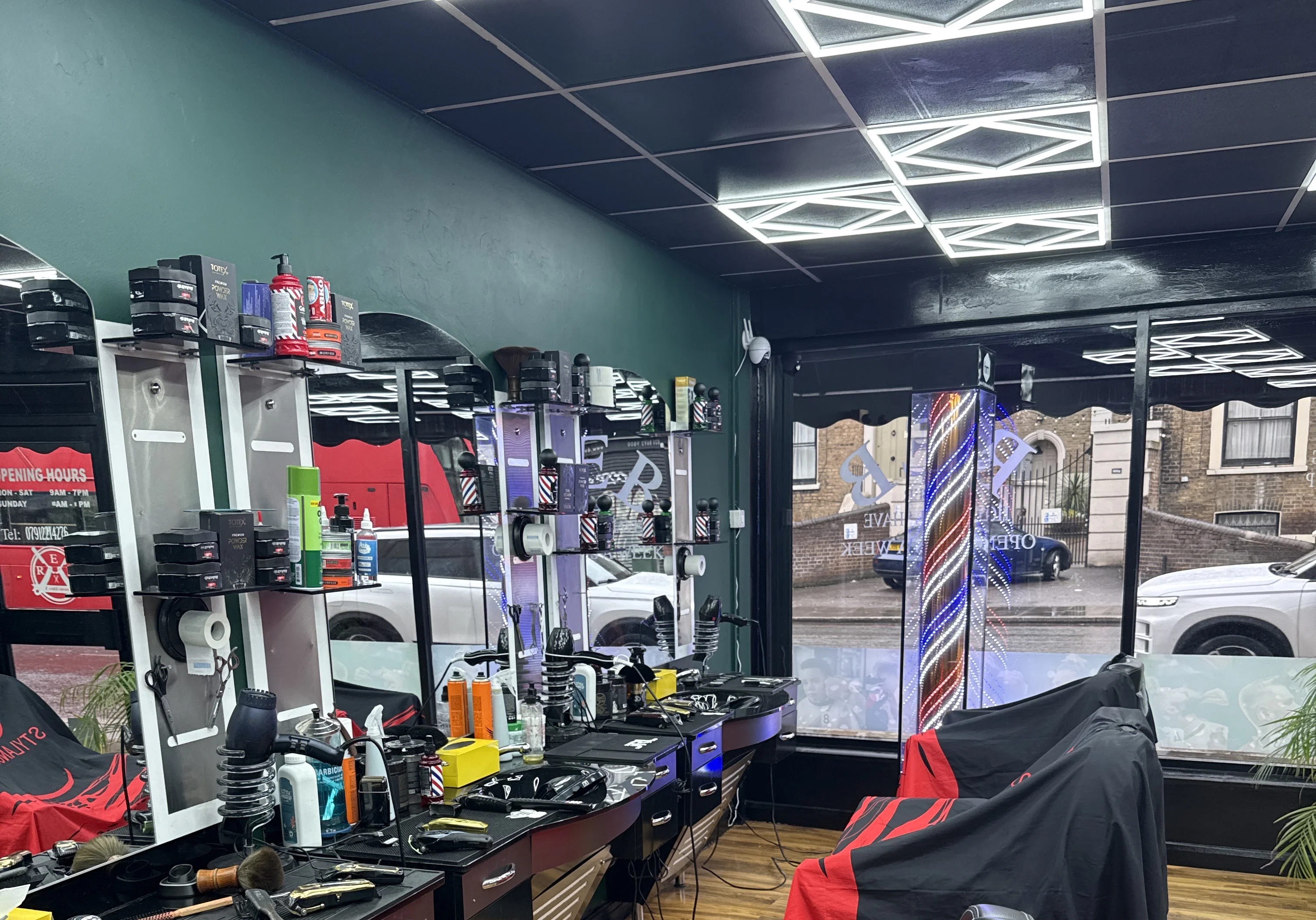 Inside Reb Barber: dark teal walls, geometric LED ceiling lights, barber stations with tools laid out, black chairs with red-and-black capes and a glowing barber-pole column