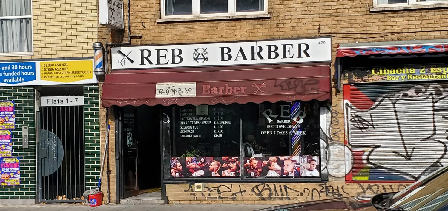 Reb Barber at 479 New Cross Road: the white REB BARBER fascia, burgundy Barber awning, barber pole, window price list and OPEN 7 DAYS A WEEK lettering