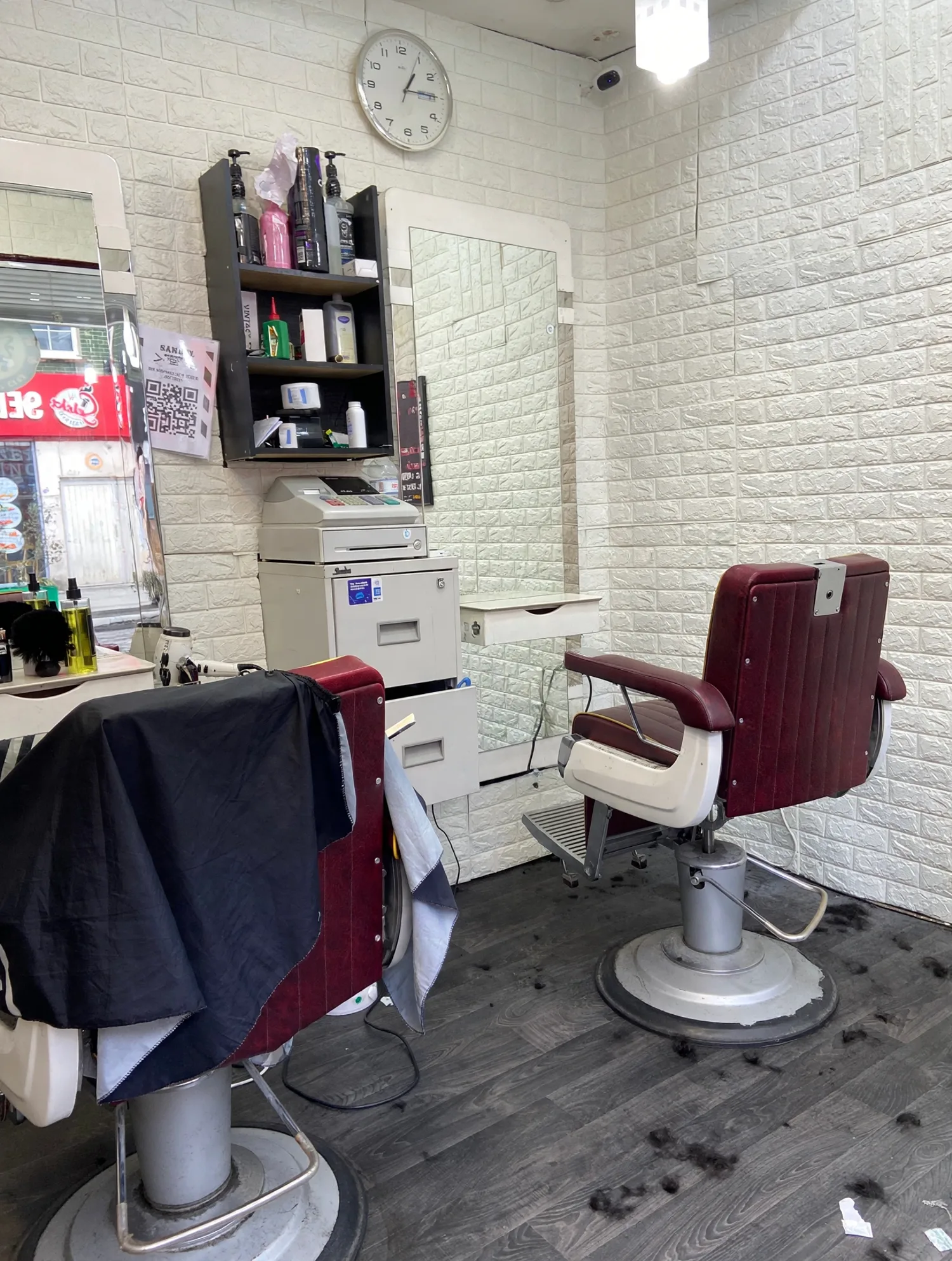 Inside Samuels Barber: two burgundy barber chairs against white brick-effect walls, with a product shelf, till and wall clock