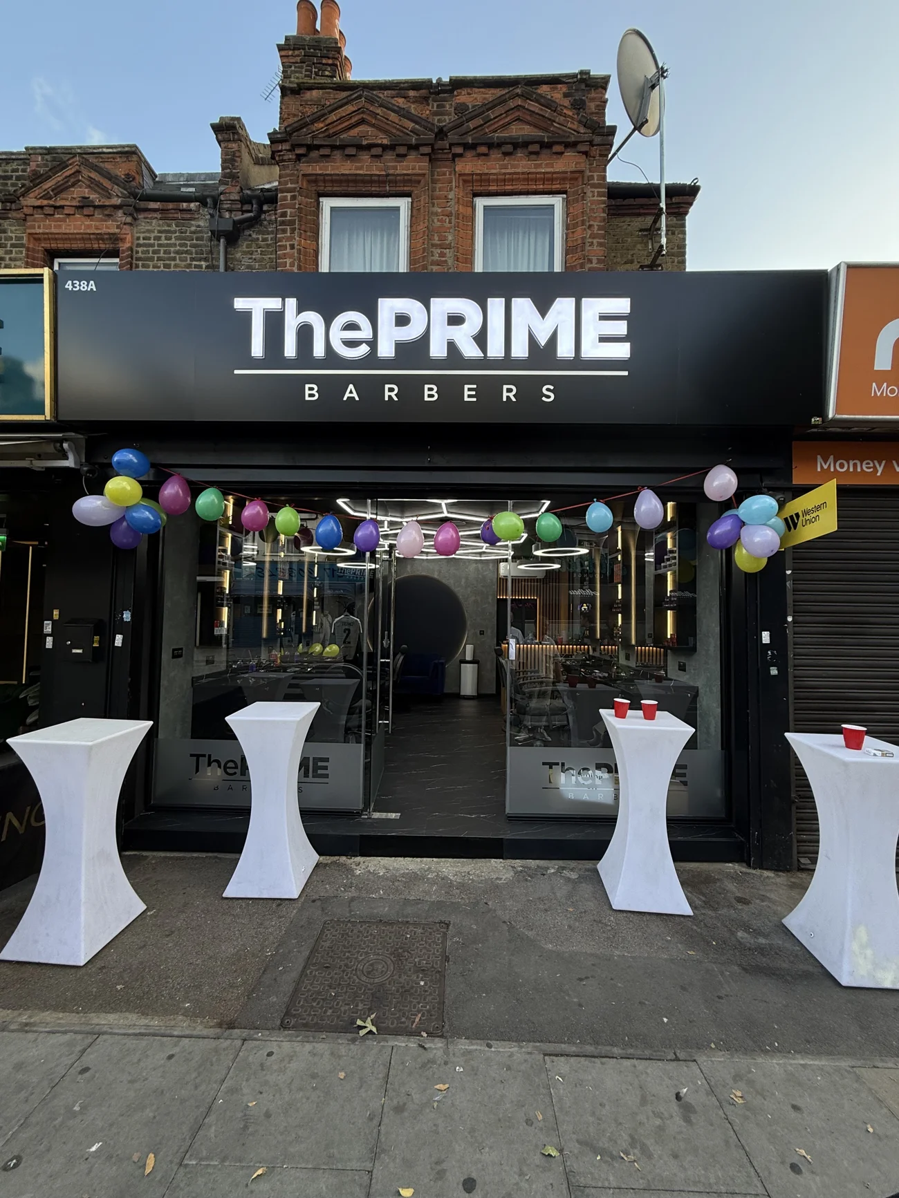 The Prime Barbers at 438A Hoe Street on its opening day: the illuminated ThePRIME BARBERS fascia, balloon garland across the window and white tables on the pavement