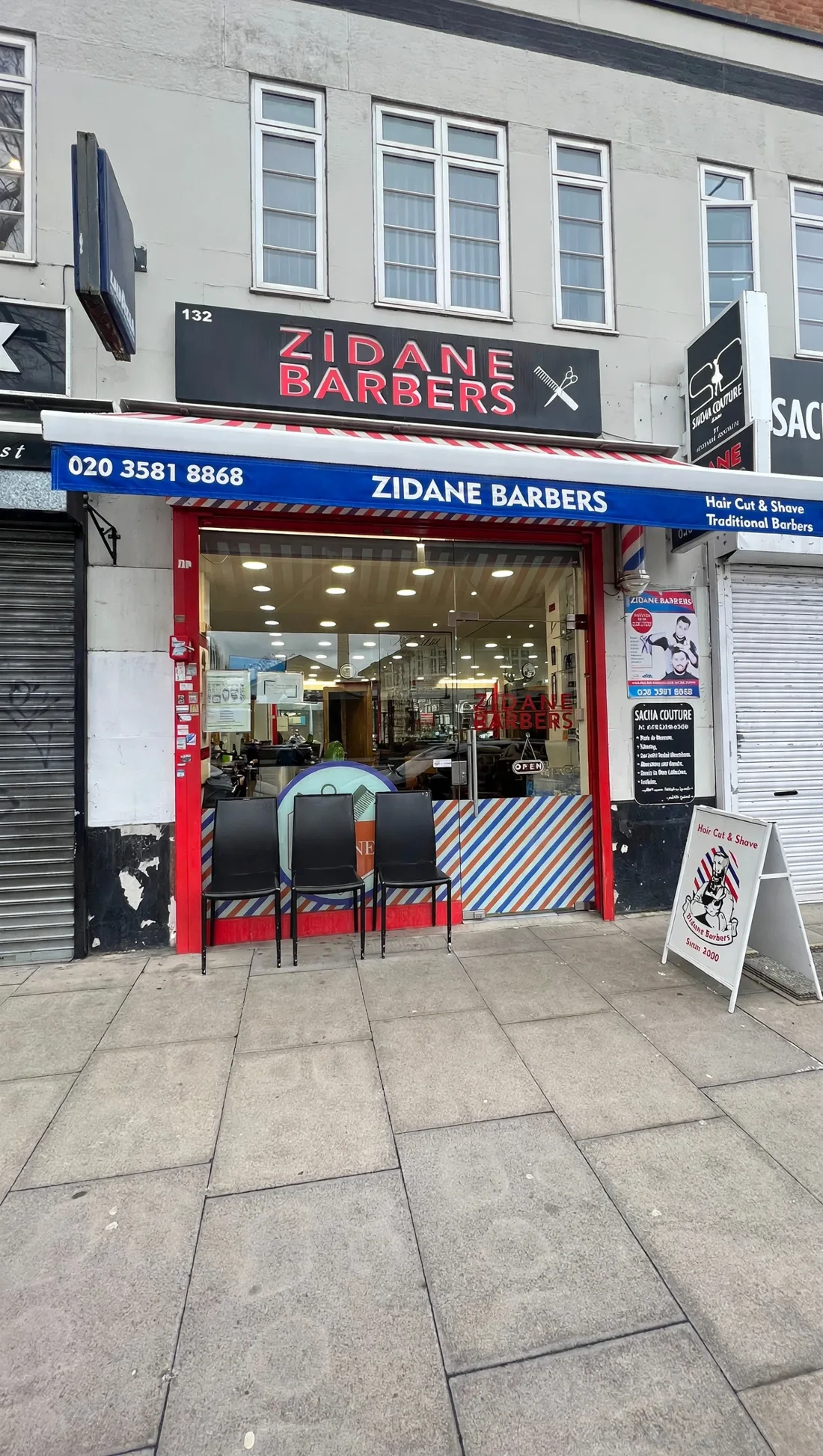 Zidane Barbers frontage on Hammersmith Road: a black fascia with red lettering, blue awning with the phone number, striped window banding and pavement chairs